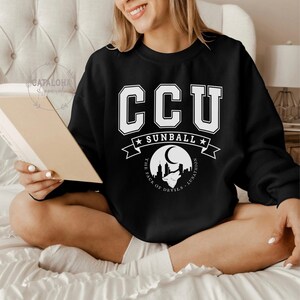 Pode incluir: Camisola preta com letras e gr&aacute;ficos brancos. A camisola apresenta as letras "CCU" em uma fonte universit&aacute;ria acima de uma faixa que diz "SUNBALL". Abaixo da faixa, h&aacute; um gr&aacute;fico de uma paisagem urbana com uma lua e o texto "THE PACK OF DEVILS - LUNATHION".