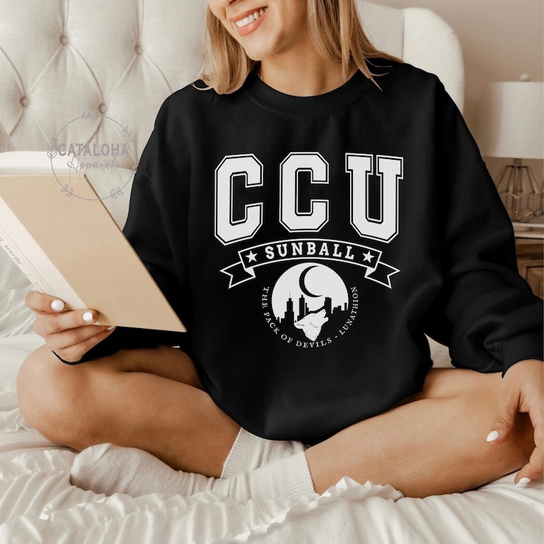 Pode incluir: Camisola preta com letras e gr&aacute;ficos brancos. A camisola apresenta as letras "CCU" em uma fonte universit&aacute;ria acima de uma faixa que diz "SUNBALL". Abaixo da faixa, h&aacute; um gr&aacute;fico de uma paisagem urbana com uma lua e o texto "THE PACK OF DEVILS - LUNATHION".