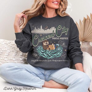 Op de afbeelding: Donkergrijze heather sweatshirt met een Crescent City Postal Service ontwerp. De afbeelding toont een otter die pakketten in het water vasthoudt, een skyline en de tekst "Sewing the Tatro River & the Bone Quarter - Friendly and efficient - Fast delivery."