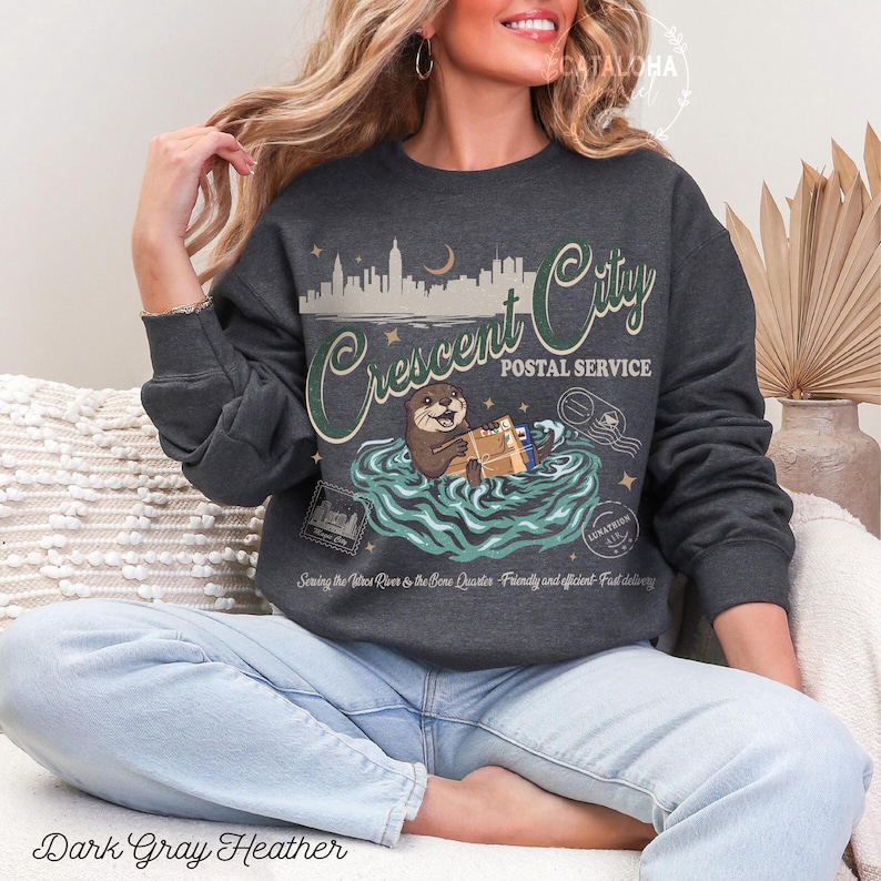 Op de afbeelding: Donkergrijze heather sweatshirt met een Crescent City Postal Service ontwerp. De afbeelding toont een otter die pakketten in het water vasthoudt, een skyline en de tekst "Sewing the Tatro River & the Bone Quarter - Friendly and efficient - Fast delivery."