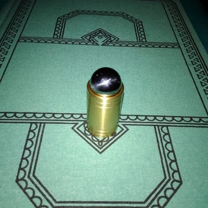 May include: A gold-colored bottle stopper with a dark, star-like stone top. The stopper is cylindrical and sits on a green surface with a black geometric pattern. The stone has a unique, multi-pointed star design.