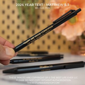 May include: Black BIC Clic pens with gold text. The pens have the text "HAPPY ARE THOSE CONSCIOUS OF THEIR SPIRITUAL NEED." and "2026 YEAR TEXT - MATTHEW 5:3." Several pens are visible. The background is blurred.