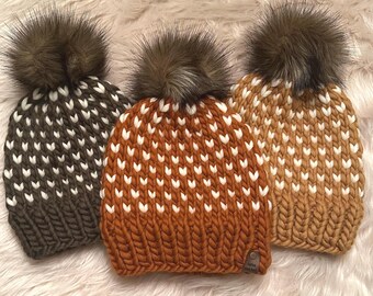 May include: Three knitted winter hats with faux fur pom-poms. The hats are in shades of brown, rust, and tan, each with a white heart-shaped pattern. The hats are displayed on a fluffy, white surface.