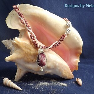 May include: A necklace made of small white and pink seashells with a large purple and white seashell pendant. The necklace is displayed on a large seashell.