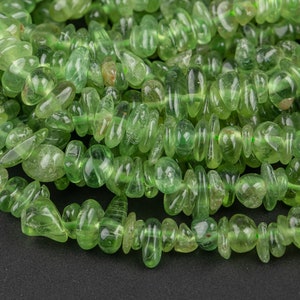 May include: A strand of green gemstone chips, also known as green aventurine, arranged in a loose pile on a black surface. The chips are smooth and polished, with a translucent appearance.