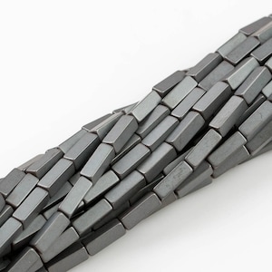 Puede incluir: Una hebra de cuentas de hematites grises, cada cuenta es un prisma rectangular con bordes ligeramente redondeados.