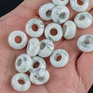 May include: A collection of white howlite donut beads with gray veining. The beads are arranged in a pile on a hand.