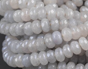May include: Close-up of multiple strands of light gray gemstone beads. The beads are faceted, with a slightly irregular shape, and strung together. The beads have a subtle, pearlescent sheen, and are arranged against a dark gray background.