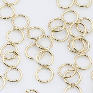 May include: A large quantity of gold jump rings, a type of jewellery finding, are scattered on a white surface. The rings are all the same size and are made of a shiny metal.