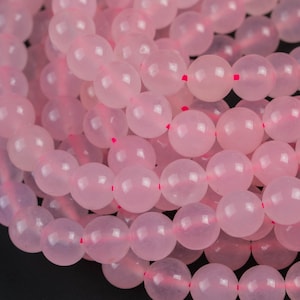 May include: Strands of light pink, round rose quartz beads. The beads are strung on a pink cord, and the image is set against a black background. The beads are smooth and polished, reflecting light.