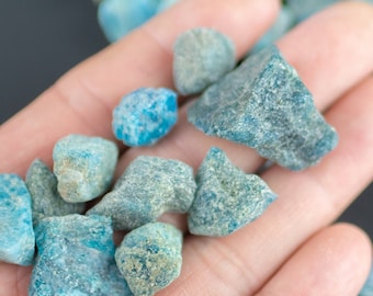 May include: A handful of small, irregularly shaped, blue-green stones. The stones vary in size and have a rough, textured surface. The stones are scattered on a dark background, with some held in a hand.