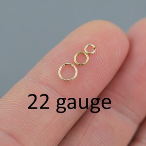 May include: Three small, gold-colored jump rings of varying sizes are displayed on a fingertip against a gray background. The text "22 gauge" is printed in black below the rings, indicating the wire thickness.
