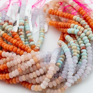 Puede incluir: Cadenas de cuentas de piedras preciosas coloridas en tonos naranja, celeste y blanco. Las cuentas están ensartadas con hilo rosa, algunas con etiquetas. Se presentan apiladas sobre un fondo blanco.