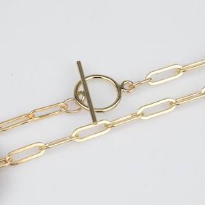 May include: A gold chain necklace with a toggle clasp. The chain is made of oval links. The necklace is lying on a white surface next to a US penny.