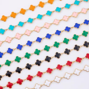 Puede incluir: Filas de collares de cadena con forma de trébol de colores en naranja, turquesa, rosa, azul, verde, negro, rojo y blanco. Las cadenas tienen un eslabón de metal dorado. Los collares se muestran en diagonal.