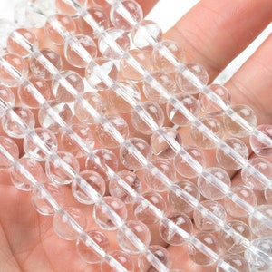 May include: Strands of clear quartz crystal beads. Each bead is round and approximately 10mm in diameter. The beads are strung together, creating a shimmering effect. The image shows the beads held in a hand against a white background.