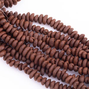 May include: Strands of brown, oval-shaped beads, likely made of wood or a similar material, are displayed against a white background. The beads are strung together, creating a textured and organic appearance. The beads are a rich, earthy brown color.