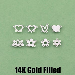 May include: Eight small silver-toned metal jewelry connectors arranged in two rows. The top row features four heart-shaped designs, while the bottom row includes a lotus and three flower shapes. Text below reads 14K Gold Filled Made in USA.