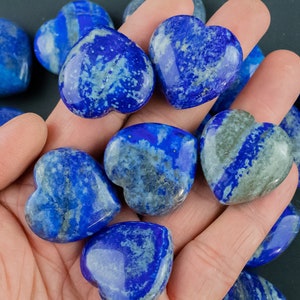 May include: A collection of heart-shaped lapis lazuli stones, each with a unique blue and white pattern. The stones are smooth and polished, and they are arranged in a hand.