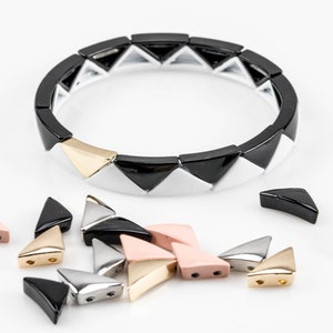 May include: A black, white and gold bracelet made of triangular ceramic beads. The bracelet is laid out flat on a white surface, with several loose beads scattered around it. The loose beads are in black, white, gold and pink.