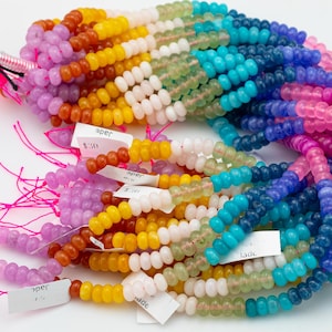 May include: A collection of colorful jade bead strands in a rainbow of hues, including pink, purple, orange, yellow, white, green, blue, and indigo. Each strand is composed of small, rounded beads. Some strands have small white tags attached.