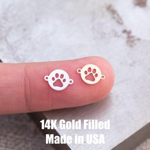 May include: Two small, round, gold-filled charms with a paw print design. The charms are on a finger, and the text "14K Gold Filled Made in USA" is below them.
