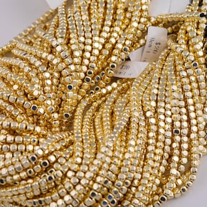 May include: Close-up of multiple strands of gold-colored beads. The beads are small, square-shaped, and strung together, creating a dense, textured appearance. The beads have a shiny, metallic finish, reflecting light. The image suggests jewelry-making supplies.