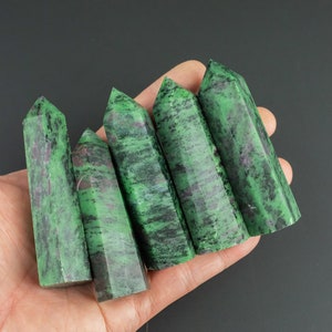 May include: Six green and black gemstone points, also known as green aventurine, are arranged in a row on a hand. The points are smooth and polished.