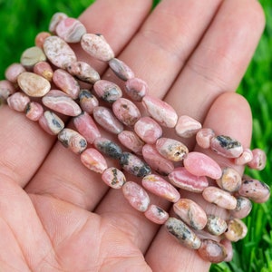 Op de afbeelding: Een streng roze en witte edelsteen kralen, met een gemarmerd patroon, in een hand. De kralen zijn ovaalvormig en hebben een gladde, gepolijste afwerking.