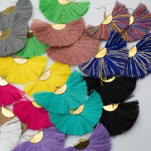 Könnte beinhalten: Eine Sammlung farbenfroher Quastenohrringe in verschiedenen Farbtönen, darunter Rosa, Gelb, Blau und Schwarz. Jeder Ohrring hat ein fächerförmiges Quastendesign mit einem goldfarbenen Metallakzent. Die Ohrringe liegen auf einer weißen Oberfläche.
