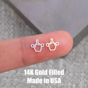 Puede incluir: Dos pendientes de oro de 14 quilates en forma de dedo medio. Los pendientes se muestran en un dedo. El texto "14K Gold Filled Made in USA" está debajo de los pendientes.