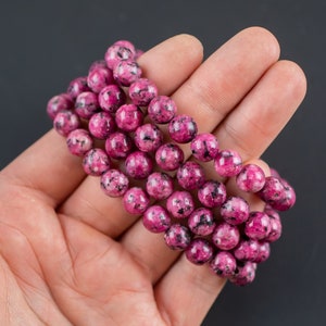May include: A pink and black speckled stone bead bracelet. The bracelet is made of multiple strands of beads and is shown in a hand.
