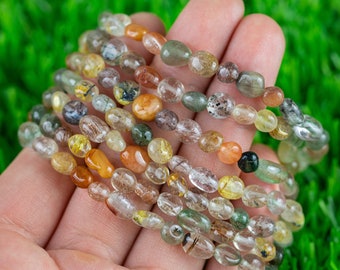 Puede incluir: Primer plano de una mano sosteniendo varios hilos de cuentas de piedras preciosas de forma ovalada. Las cuentas son translúcidas y tienen una variedad de colores, incluyendo transparente, marrón, verde y amarillo. Las cuentas están dispuestas en una pila, con la cuenta superior de cada hilo visible.