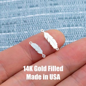Puede incluir: Dos pequeños dijes dorados en forma de pluma con agujeros para hacer joyas. Los dijes se muestran sobre un fondo azul claro. El texto en la parte inferior dice "14K Gold Filled Made in USA."