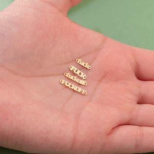 Puede incluir: Cuatro pequeños dijes de metal de color dorado dispuestos en una fila vertical. Cada dije presenta una palabra diferente recortada en una fuente estilizada: "fuck", "fucks", "fucker" y "fucko". Cada pieza tiene un pequeño aro en los extremos.
