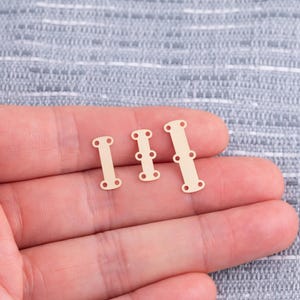 May include: Three small, gold-colored metal clasps in varying sizes. Each clasp has a rectangular shape with rounded ends and small holes for attaching to fabric or other materials. The clasps are shown on a light gray background.