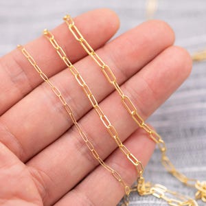 May include: Close-up of several gold-colored paperclip chain necklaces. The chains vary in size, with the links ranging from small to large. The necklaces are displayed on a light-colored surface.