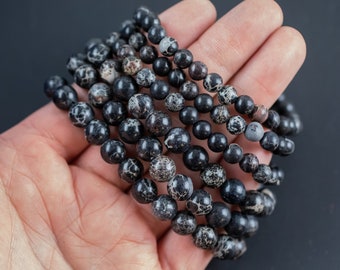 May include: Several black and gray beaded bracelets of varying sizes are held in a hand against a dark background. The beads have a speckled pattern, and the bracelets appear to be made of natural stone.