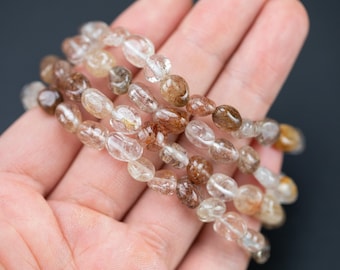 Op de afbeelding: Een streng bruine en heldere ovale kralen met een metallic glans. De kralen zijn geregen op een heldere elastische koord.