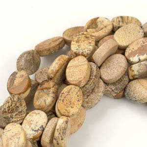 May include: A strand of oval-shaped, brown and beige, natural stone beads. The beads have a mottled pattern and are strung together to create a necklace or bracelet.