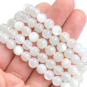 May include: A strand of white moonstone beads, each bead is round and has a smooth, polished surface. The beads are strung together on a clear thread.