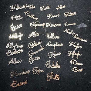 May include: A collection of gold-colored metal name tags with various names written in cursive script. The names include Kohenoot Fitido, Karlo, Aloo, Hitdo, Castano, Finno, Zedeo, Jasco, Elway Kasoro, Emma, Mellafo, Allo, Valero, Nicole Lawsono, Lily Anne, Cabine, Boulevo, Cavannabo, Achers, Jace, Rilano, Klacho, Landero, Fjoo, Judabo, Draw, Case, and Eatoire.