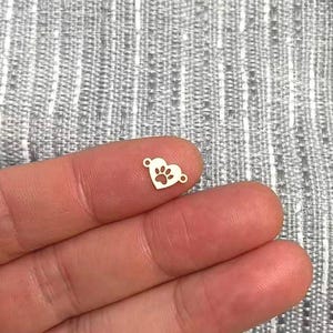 Puede incluir: Un pequeño dije plateado en forma de corazón con un diseño de huella de pata. El dije tiene dos pequeños bucles a cada lado para sujetarlo a una pulsera o collar. El dije está sostenido en la mano.
