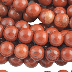 May include: A strand of red jasper beads, each bead is round and smooth with a polished finish. The beads are arranged in a loose pile, showing the natural variations in the stone's color and pattern.