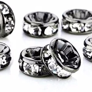 May include: A collection of six round, gunmetal-toned metal spacer beads. Each bead features a central hole and is embedded with a row of clear, faceted crystals around the outer circumference, creating a reflective and textured appearance.