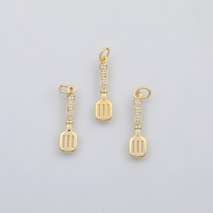May include: Three gold-coloured spatula charms with a slotted design. The handles are encrusted with small, clear gemstones. Each charm has a small loop at the top for attaching to a necklace or bracelet. The charms are on a white background.