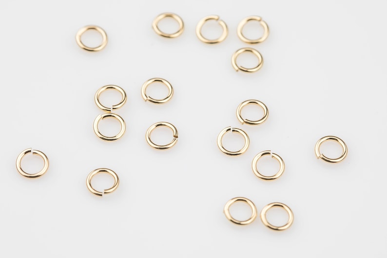 May include: A collection of small, gold-colored jump rings scattered on a white surface. Some rings are open, while others are closed. These jewelry findings are used for connecting jewelry components.