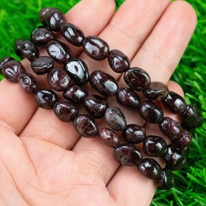 May include: A strand of dark red gemstone beads, likely garnet, strung together in a long, oval shape. The beads are smooth and polished, with a slight sheen.