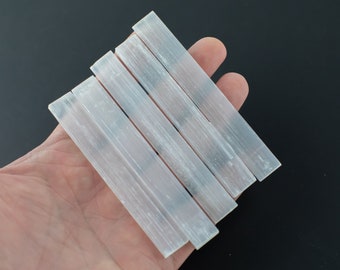 Könnte beinhalten: Sechs weiße Selenit-Stäbe, auch bekannt als Selenit-Stäbe, liegen übereinander in einer Hand. Die Stäbe sind glatt und haben ein natürliches, weißes, kristallines Aussehen.