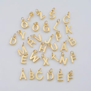 May include: A collection of gold-colored alphabet letter charms, each with a small loop for attaching to jewelry. The letters are in a variety of fonts and sizes, and are scattered across a light background. These charms are ideal for personalized jewelry.